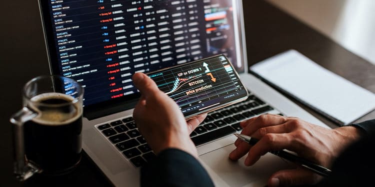Hombre evaluando el mercado de las criptomonedas en la pantalla de un teléfono y en la computadora. Fuente: avanti_photo / elements.envato.com