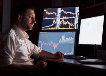 Trader en habitación oscura con gráficos de mercados en pantallas. Fuente: mstandret / elements.envato.com